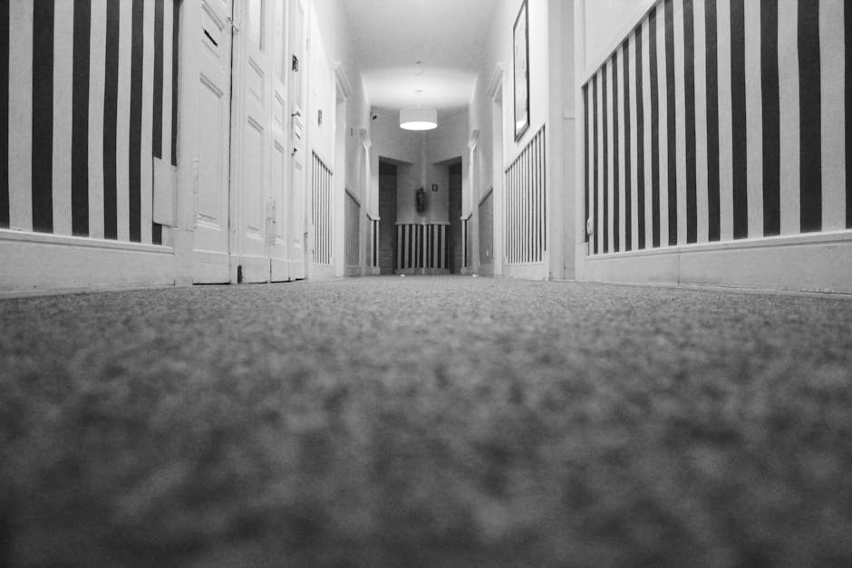 A person wearing light-colored overalls is performing surface cleaning using a yellow and black vacuum cleaner on a grey carpeted floor inside a room with dark walls and a white ceiling-mounted projec