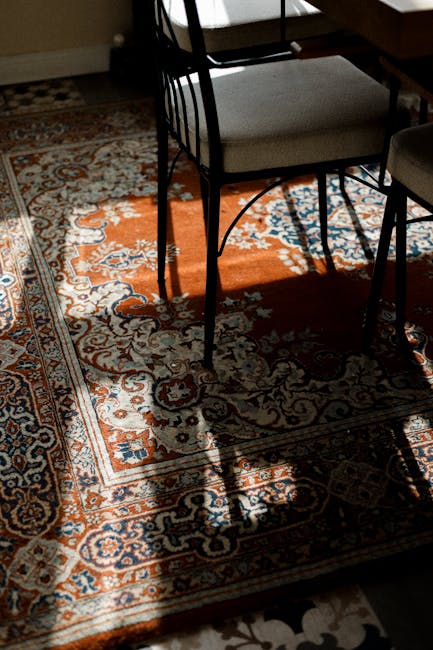 Close-up view of a richly patterned area rug with intricate floral and geometric designs in shades of orange, blue, and cream, placed on a wooden floor in a well-lit dining area. The rug appears clean